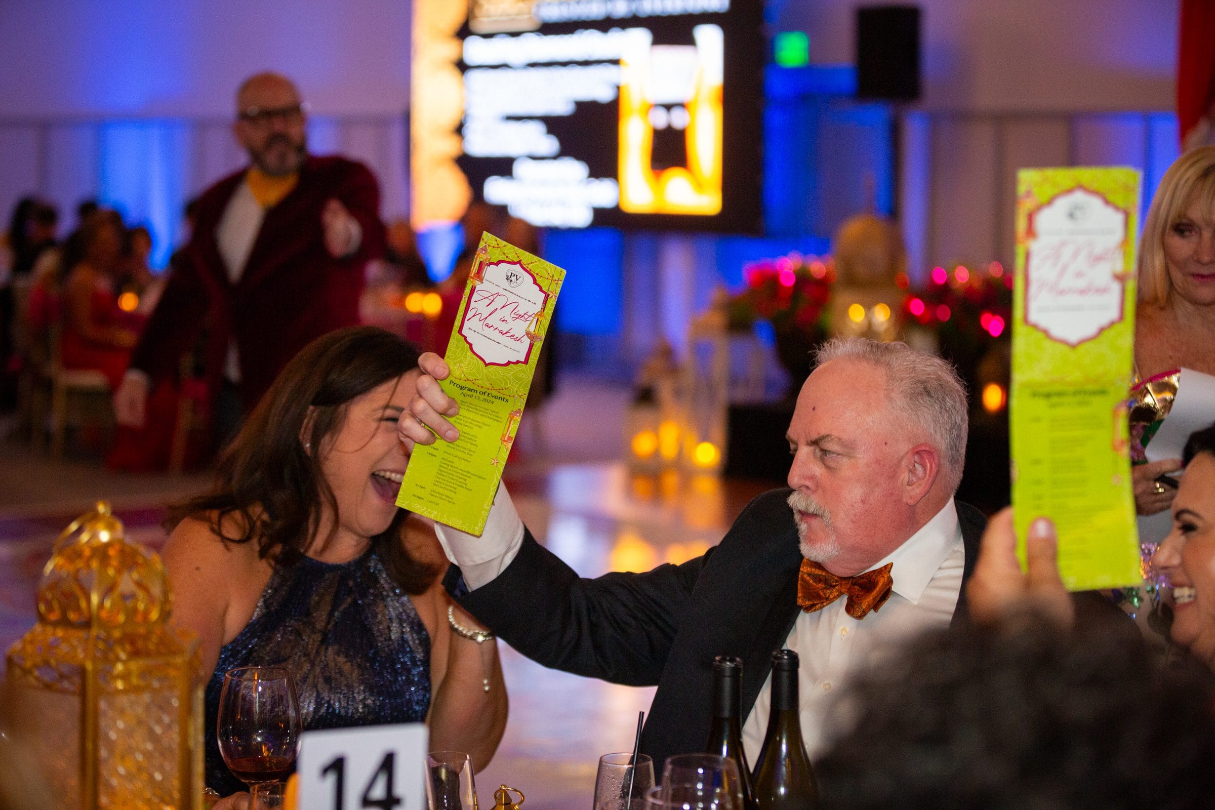 Guests laugh and raise event program as auctioneer entertains nearby.